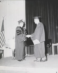 Commencement--Dr. Harden and Bruce Anderson Jan. 1961: Two Men in Graduation Gowns Shaking Hands on Stage